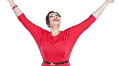 Eine lächelnde Frau in einem roten Kleid hebt die Arme in die Luft, fröhlich und selbstbewusst, vor einem weißen Hintergrund.