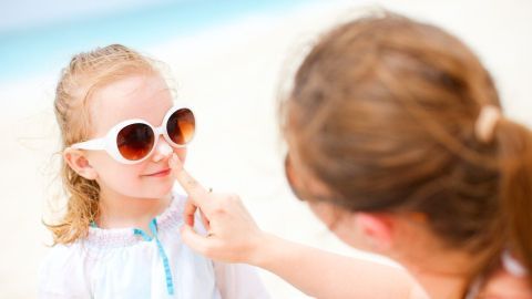 Ein Mädchen mit Sonnenbrille sitzt am Strand, während eine andere Person ihr sanft Sonnencreme aufträgt.