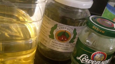 Ein Glas mit Etikett steht neben einer Flasche Öl und einem weiteren Glas in einer hellen Umgebung.