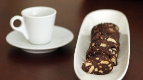 Schokoladenwurst liegt auf einer weißen Platte neben einer kleinen Tasse Kaffee, auf einem Holzuntergrund.
