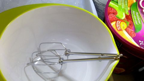 Eine Schüssel mit einem Handmixer steht bereit, um schnell Sahne zu schlagen, während bunte Eisbehälter im Hintergrund stehen.
