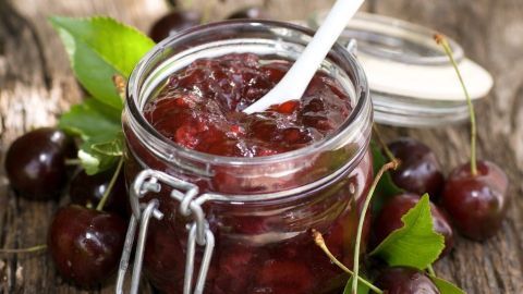 Ein Glas Sauerkirsch-Schokoladen-Marmelade steht auf einem rustikalen Holztisch, umgeben von frischen Kirschen und grünen Blättern.