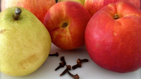 Bunte Äpfel und eine Birne liegen zusammen mit Gewürznelken auf einem weißen Tisch, ideal zur Fruchtfliegenabwehr.