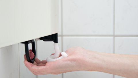 Eine Hand drückt einen Deodorant-Spender in einem Badezimmer, während in der Umgebung weiße Fliesen sichtbar sind.