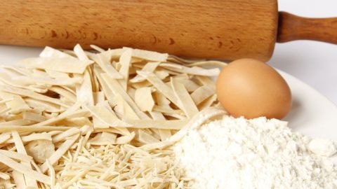 Auf einem weißen Tisch liegt ein Nudelholz neben frischen Teigstreifen, einem Ei und Mehl, bereit zum Nudeln selber machen.