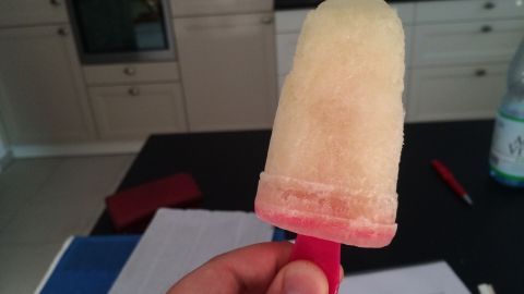 Ein Radler-Eis am roten Stiel wird in einer modernen Küche gehalten; im Hintergrund sind ein Backofen und ein Tisch zu sehen.