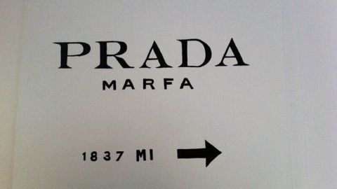 Ein Schild mit der Aufschrift „PRADA MARFA“ zeigt, dass man 1837 Meilen in diese Richtung fährt, auf weißem Hintergrund.