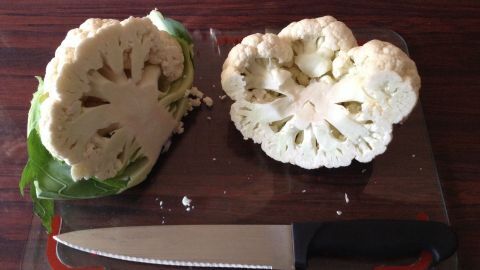 Frische Blumenkohlhälften liegen auf einem Schneidebrett, ein scharfes Messer daneben, bereit zum Rohbraten.