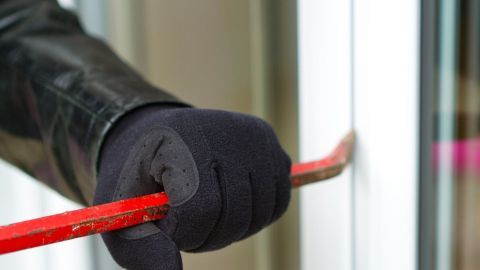 Eine Hand mit schwarzer Handschuh zieht einen roten Brecheisen in eine Tür, um Einbruchschutz zu umgehen.