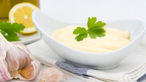 Eine feine helle Grundsoße in einer Schüssel, umgeben von Knoblauch und einer Zitronenscheibe auf einem Tisch.