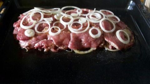 Rohes mariniertes Gyrosfleisch liegt auf einem Backblech, garniert mit geschnittenen Zwiebeln, bereit zum Braten im Ofen.