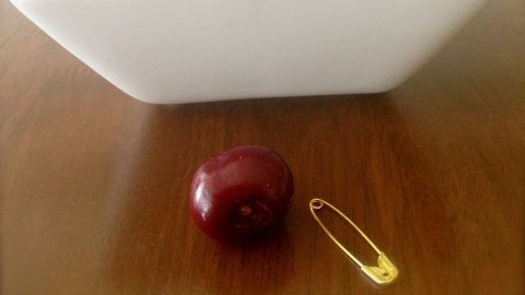 Eine Schüssel mit Kirschen steht auf einem Holz Tisch, während eine Kirsche und eine Sicherheitsnadel daneben liegen.