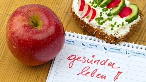 Ein roter Apfel liegt neben einem belegten Brot mit Gemüse, während ein Kalender den Text "gesünder leben!" zeigt.