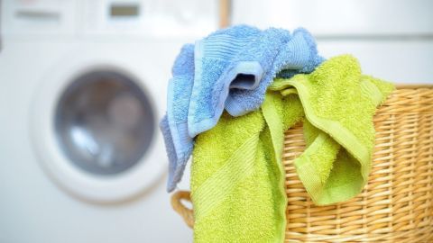 Bunte Handtücher liegen auf einem Wäschekorb, während im Hintergrund eine Waschmaschine bereitsteht, um sie richtig zu waschen.
