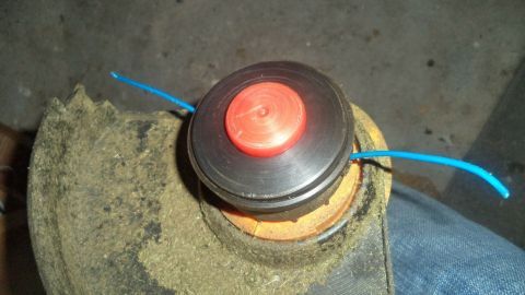 Ein Rasentrimmerkopf mit einer rotierenden Spule und blauen Schnüren ist in einer Werkstattumgebung montiert.