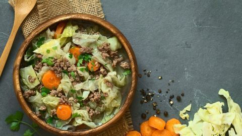 Ein Holzschüssel enthält herzhaften Kohlpudding mit Rinderhackfleisch und Karotten, umgeben von Gewürzen und frischem Gemüse.