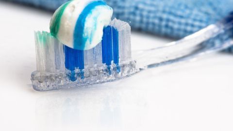 Zahnpasta liegt in bunten Streifen auf einer nassen Zahnbürste, die auf einem weißen Untergrund neben einem blauen Tuch liegt.