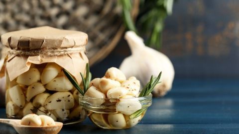 Ein Glas mit eingelegtem Knoblauch steht auf einem Holzuntergrund, daneben frischer Knoblauch und ein Löffel mit Knoblauchzehen.