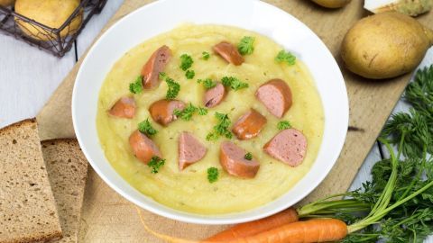 Eine Schüssel Eintopf mit Würstchen und frischen Zutaten wie Kartoffeln und Karotten auf einem Tisch, umgeben von Brot.