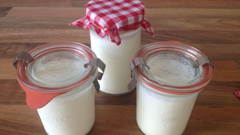 Selbstgemachter Joghurt füllt drei Gläser auf einer Holzoberfläche, zwei mit Deckel und eins mit rot-weißem Stoffbezug.