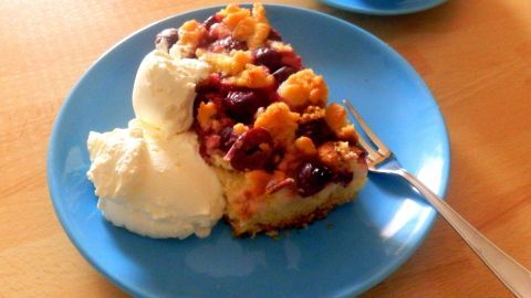 Ein Stück Sauerkirsch-Käsekuchen liegt auf einem blauen Teller, daneben ein Klecks Vanilleeis, auf einem Holztisch.
