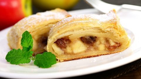 Auf einem Teller liegt ein Stück Apfelstrudel, gefüllt mit Äpfeln, auf einem Tisch mit einem grünen Apfel daneben.