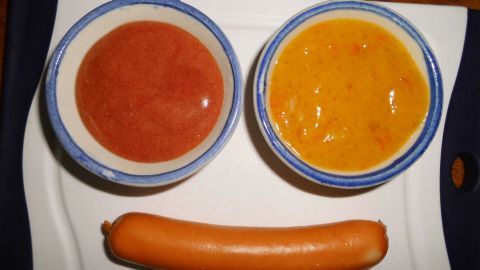 Auf einem Teller liegen zwei Schalen mit Senf, einer rot, einer gelb, über einer Wurst, die wie ein Lächeln aussieht.