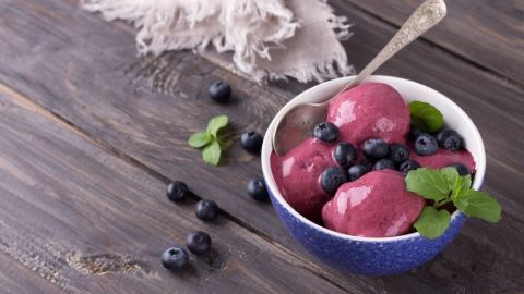 In einer Schüssel liegt fruchtiges Eis, garniert mit frischen Heidelbeeren und Minzblättern, auf einem rustikalen Holz Tisch.