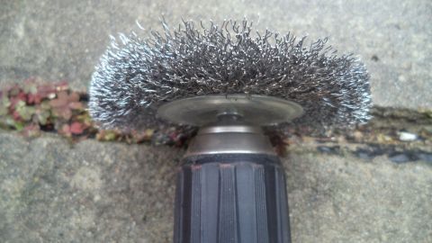 Eine Drahtbürste liegt auf einem Betonboden und entfernt Unkraut aus Fugen zwischen den Platten.