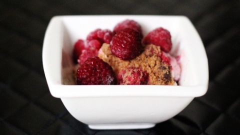 Ein Dessert mit Himbeeren und Kekskrümeln in einer weißen Schüssel, platziert auf einer dunklen Unterlage.