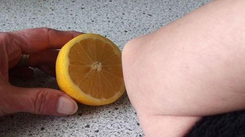 Hornhaut an Ellbogen oder Knie entfernen mit Zitrone. Eine Hand hält eine aufgeschnittene Zitrone neben einem Arm, um Hornhaut zu entfernen. Die Oberfläche ist eine helle Arbeitsplatte.
