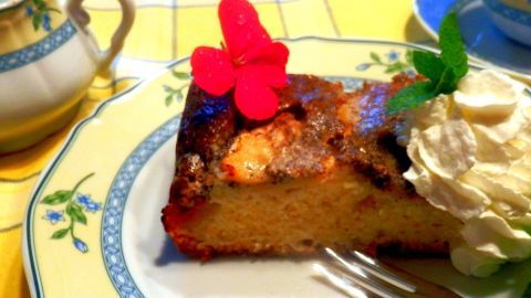 Ein St&uuml;ck Mohn-Apfelkuchen auf floralem Teller mit Sahne und Minze garniert, neben Porzellantasse auf gelbem Tischdeckenmuster.