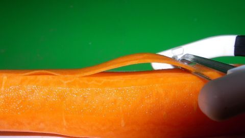 Eine Karotte wird mit einem Sparschäler geschält, während sie auf einer flachen Oberfläche liegt. Der Hintergrund ist grün.