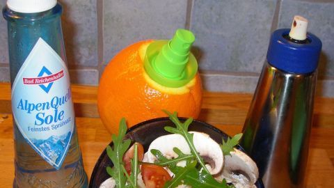 Ein Sprühbehälter für Salz und ein weiterer für Gewürze stehen neben einer Schüssel mit frischem Salat und einer Orange.