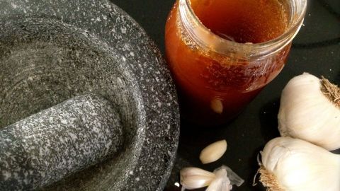 Ein Mörser liegt neben einem Glas Knoblauch-Honig-Paste und frischen Knoblauchzehen auf einem dunklen Untergrund.