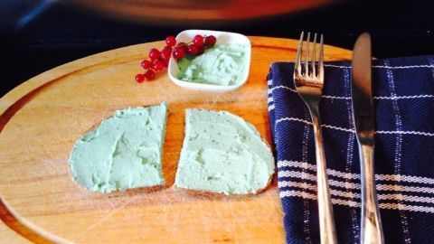 Auf einem Holzbrett liegen zwei Scheiben Brot mit grünem Oliven-Brotaufstrich, daneben ein Schälchen und Besteck.