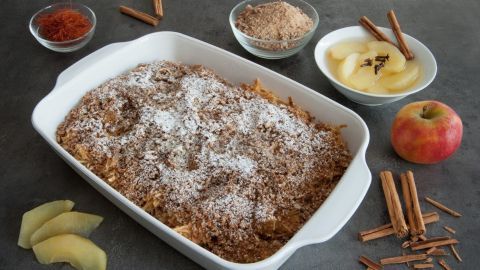 Ein Apfelauflauf mit Roggenbrot ist goldbraun gebacken, bestreut mit Puderzucker, umgeben von Äpfeln und Gewürzen.
