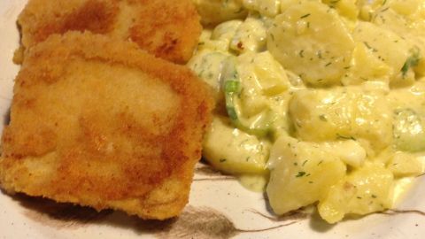 Sellerieschnitzel liegen auf einem Teller neben einem Kartoffelsalat, der mit frischen Kräutern verfeinert ist, auf grünem Platzdeckchen.