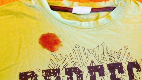 Ein T-Shirt liegt auf einem Holzfußboden, sichtbar mit einem Himbeerfleck. Der Fleck ist rot und rund, auf dem Stoff.
