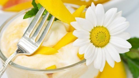 Ein glasiges Mango-Kokos-Dessert ist dekoriert mit Ananasstücken und Gänseblümchen, umgeben von einer Schale frischer Mangostücke.