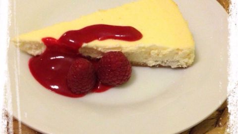Ein Stück Himbeer Cheesecake liegt auf einem weißen Teller, garniert mit Himbeeren und fruchtiger Sauce, auf einem geflochtenen Untersetzer.