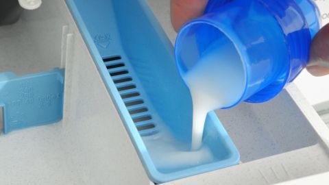 Ein blauer Messbecher wird mit Weichspüler gefüllt und über eine offene Schublade einer Waschmaschine gehalten.