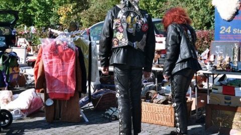 Verkäufer in Lederjacken durchsuchen bunt gemischte Flohmarktstände in einem offenen, grünen Park bei sonnigem Wetter.