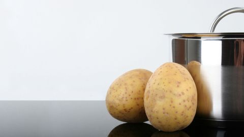 Ein glänzender Edelstahltopf steht auf einer glatten Oberfläche, neben ihm liegen zwei Kartoffeln.
