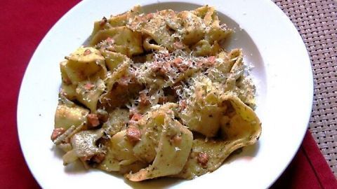 Breite Bandnudeln liegen auf einem weißen Teller, garniert mit Walnuss Pesto und vegetarischem Schinken, serviert auf einem Tisch.