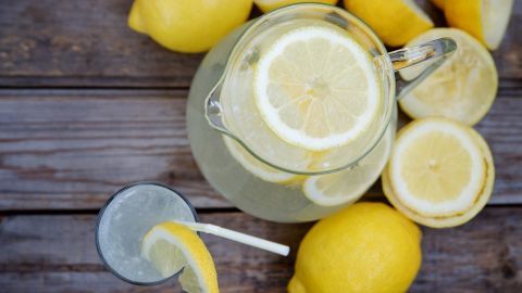 Ein Glas Limonade steht neben einem Krug gefüllt mit Limonade und frischen Zitronen auf einem Holztisch.
