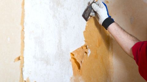 Eine Hand entfernt mit einem Spachtel alte Tapete von einer Wand in einem hellen Raum, um die Oberfläche vorzubereiten.