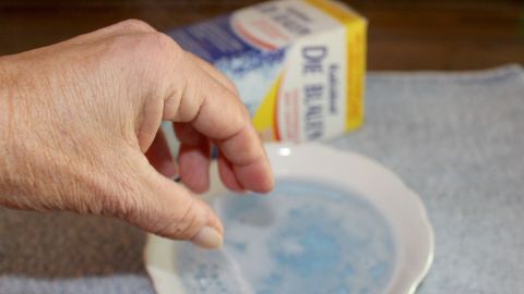 Hand taucht Gebissreiniger in eine Schale. Im Hintergrund eine Packung mit Aufschrift "Die Blauen" auf einem Tuch.