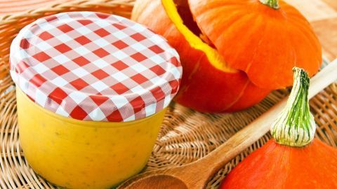 Eine Glas mit Kürbiscremesuppe steht auf einem Korb, umgeben von frischen Kürbissen und einem Holzlöffel.