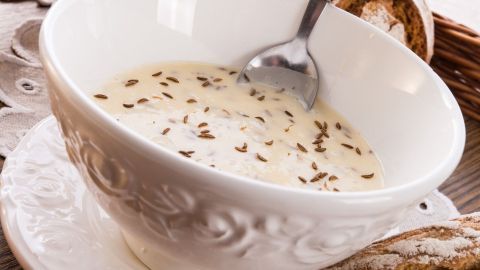 In einer dekorativen Schüssel liegt cremiger Kochkäse, garniert mit Kümmel, daneben frisches Brot im Hintergrund.